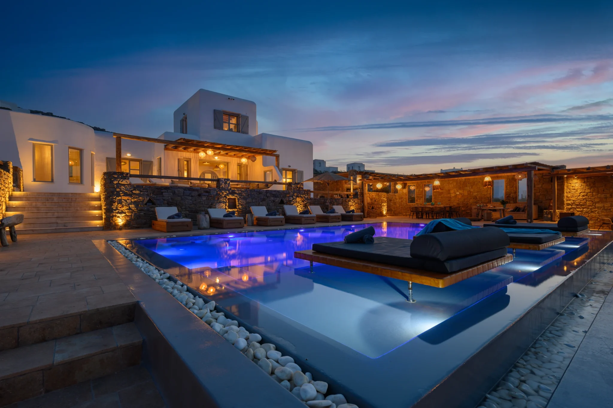 White villa and warmly lit stone pergolas beside an illuminated blue swimming pool beneath a pastel evening sky at Villa Ostraco in Mykonos.