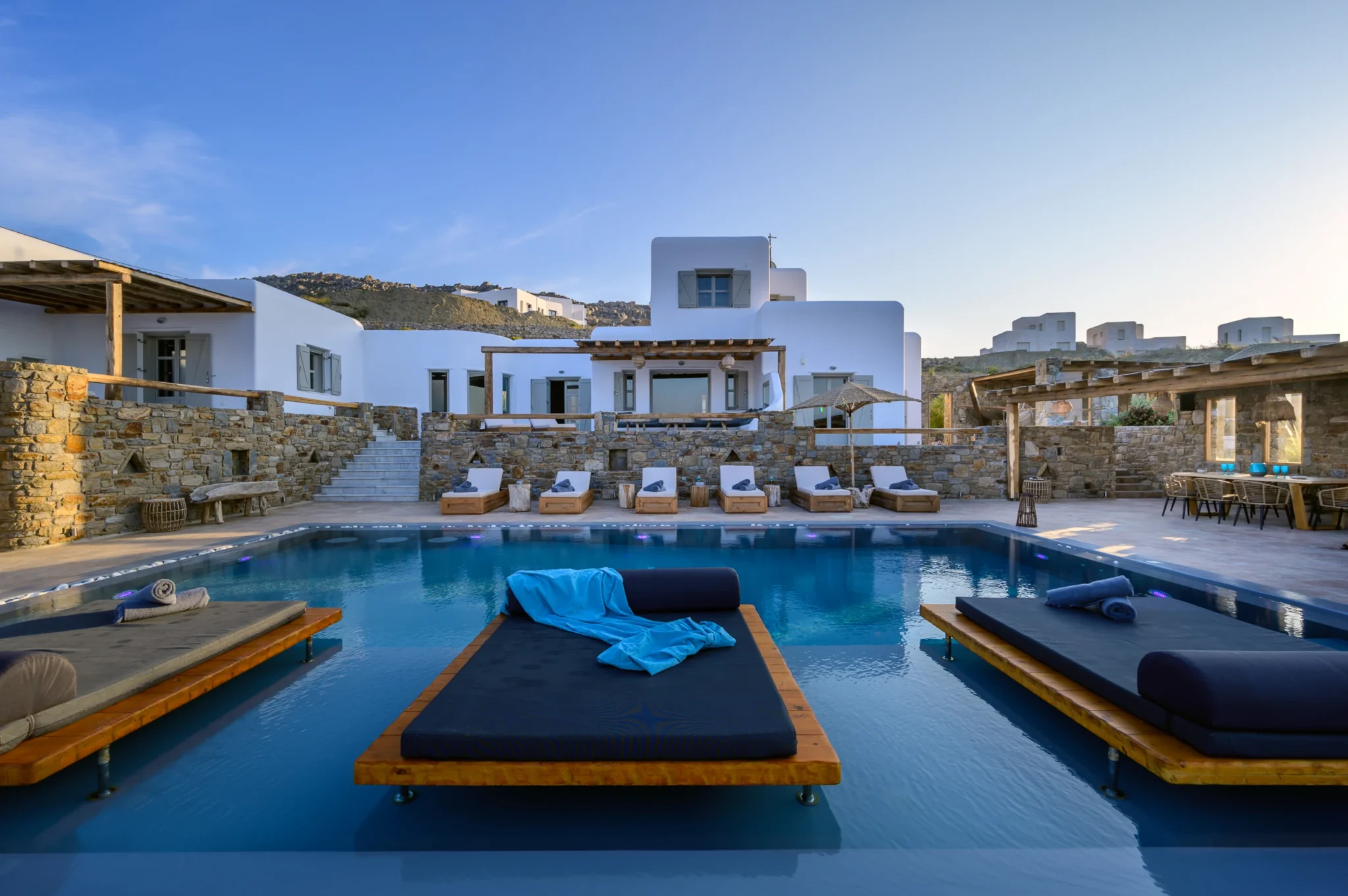 White Cycladic villa and stone terraces seen across the swimming pool, with three cushioned platform daybeds in the foreground at Villa Ostraco in Mykonos.