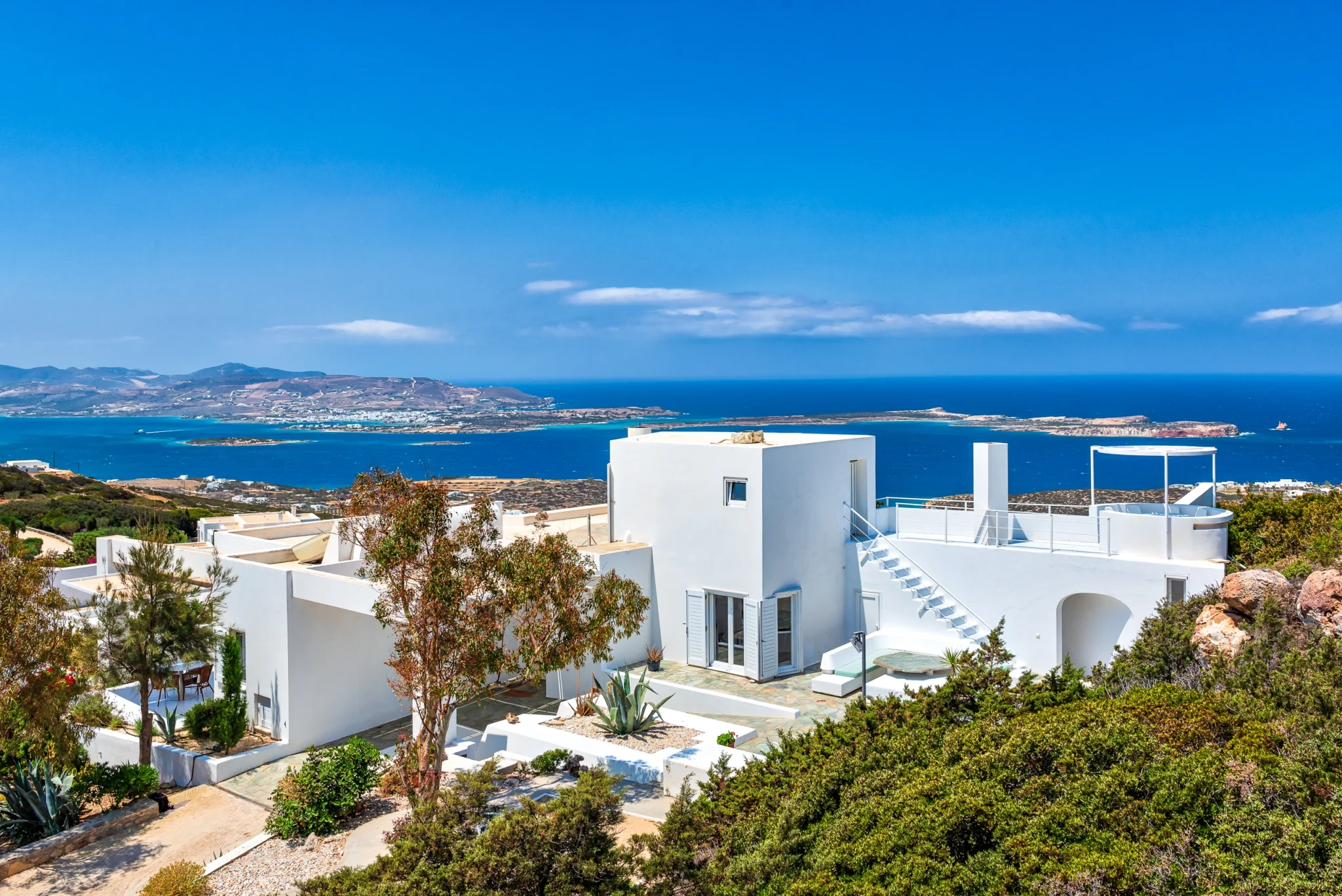 Elevated view of Villa Pastorale in Paros, showing white cubic volumes, external stairs and planted stone terraces overlooking the sea and islands.