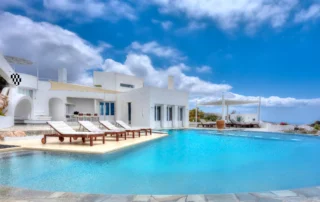 White Cycladic architecture of Villa Pastorale in Paros beside a curved turquoise pool, with wooden sun loungers, a shaded courtyard and a white pergola.