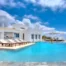 White Cycladic architecture of Villa Pastorale in Paros beside a curved turquoise pool, with wooden sun loungers, a shaded courtyard and a white pergola.