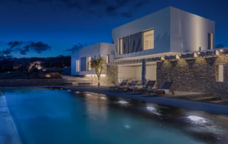 Illuminated white and stone villa reflected in the pool at blue hour at Villa Seafront, Paros.