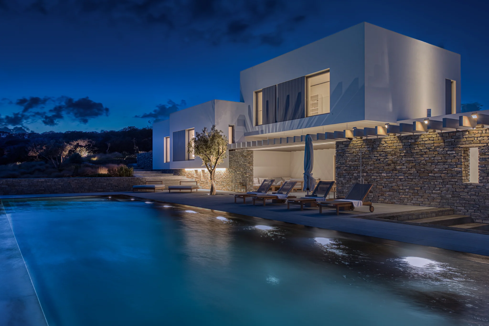 Illuminated white and stone villa reflected in the pool at blue hour at Villa Seafront, Paros.