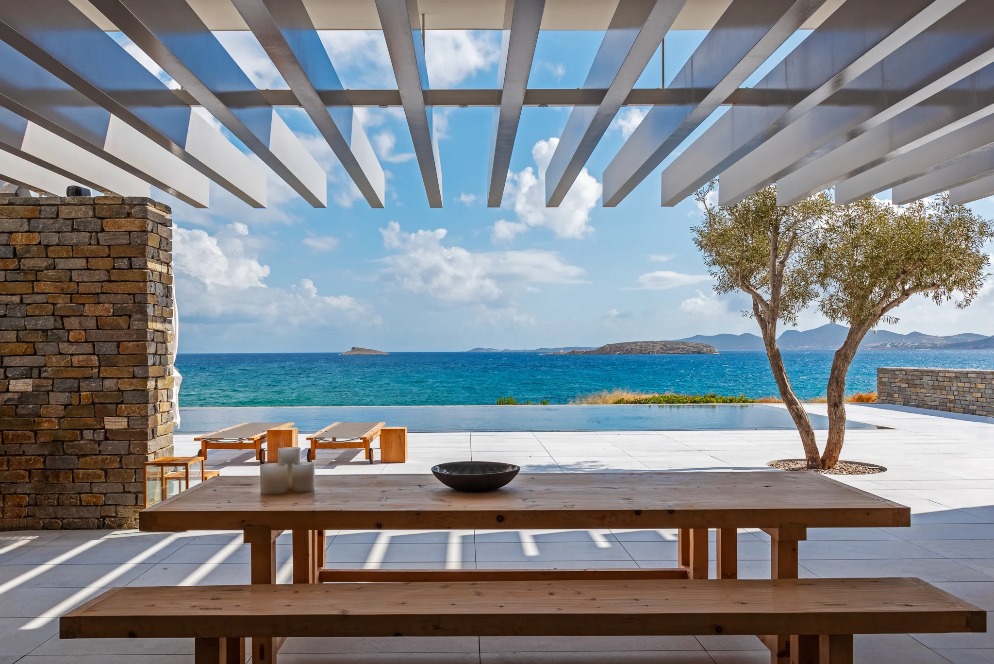 Timber dining table beneath a pergola overlooking the pool and sea at Villa Seafront, Paros.