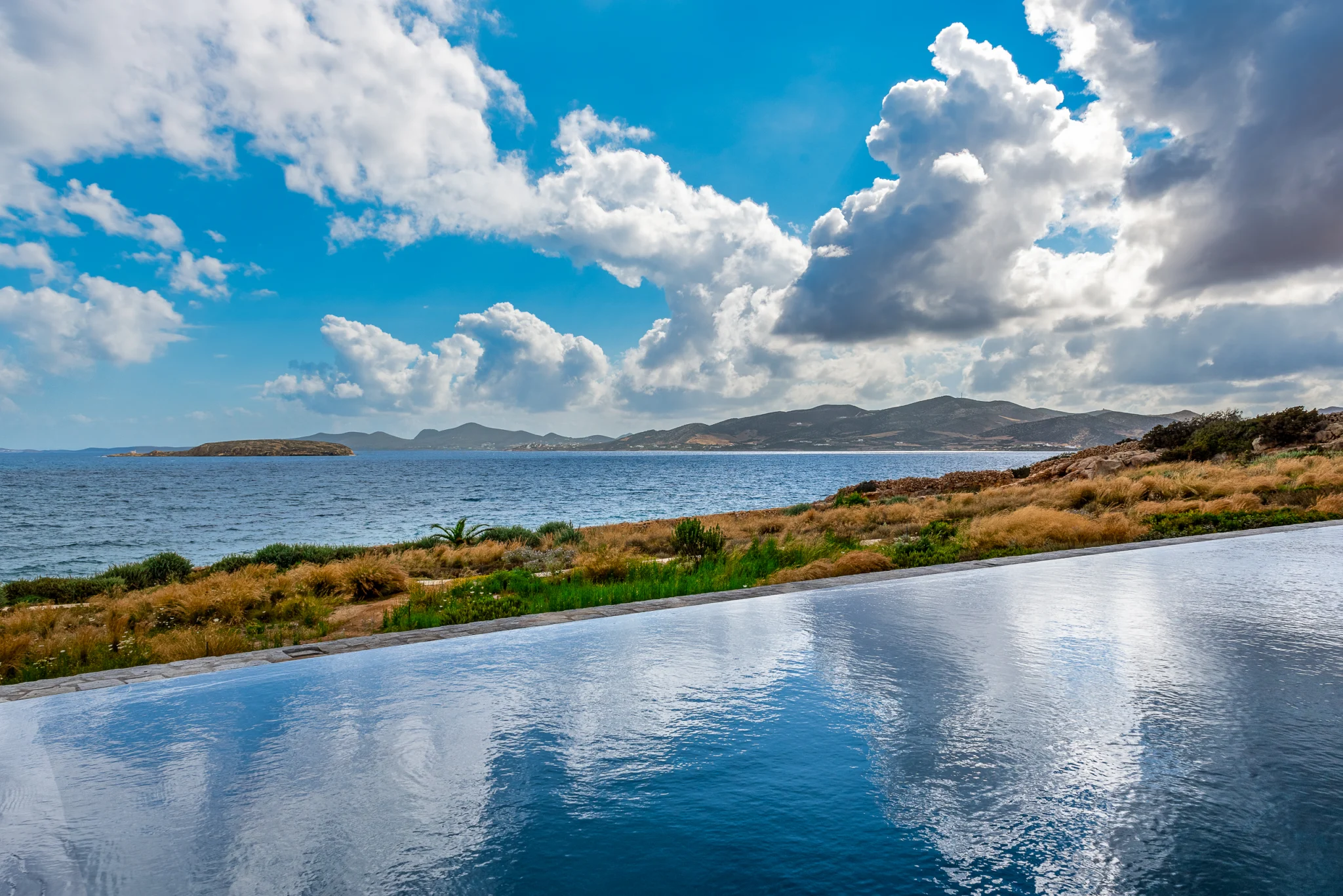 Infinity pool reflecting clouds beside coastal planting and the sea at Villa Seafront, Paros.