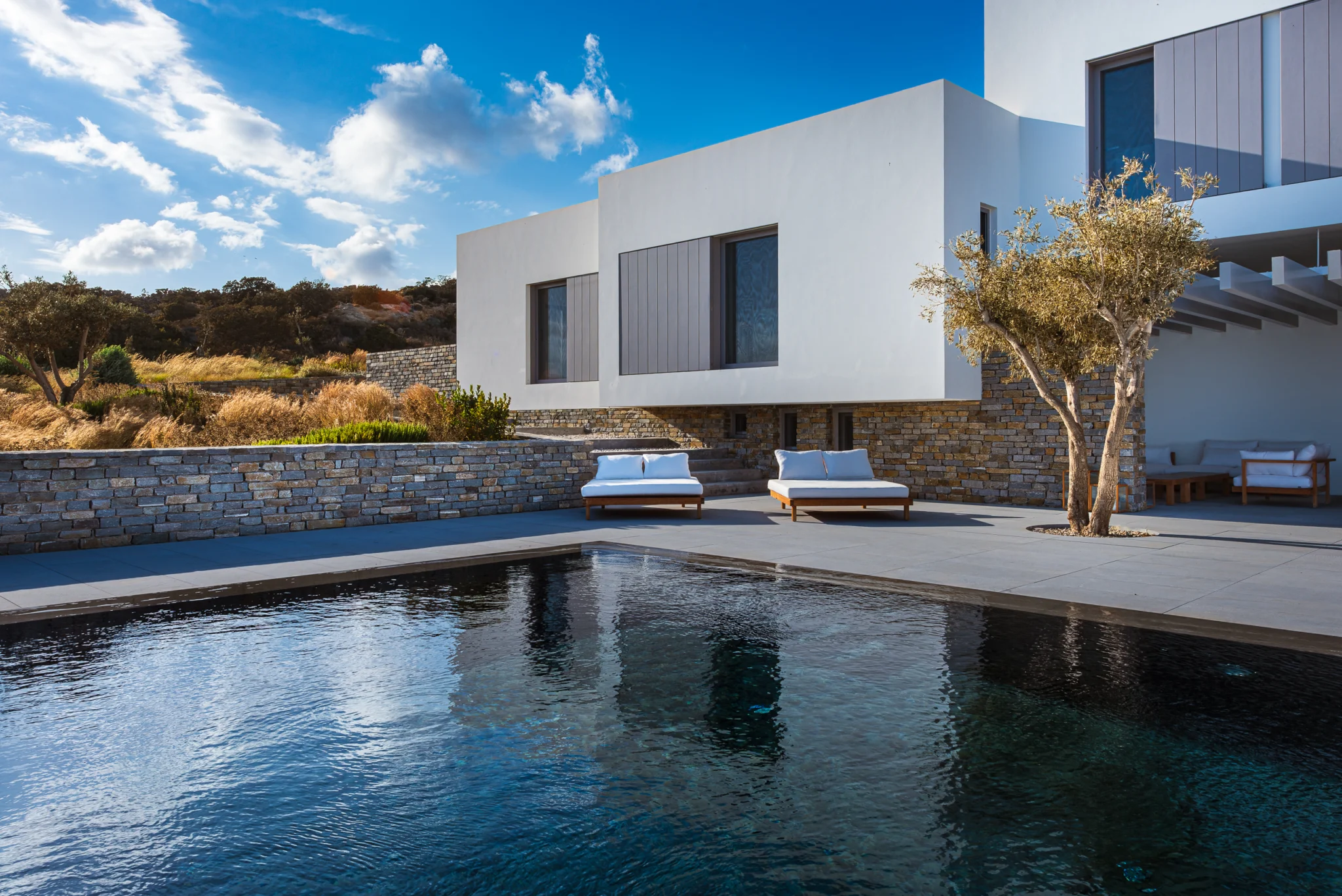 Pool terrace beside a projecting white volume and an olive tree at Villa Seafront, Paros.