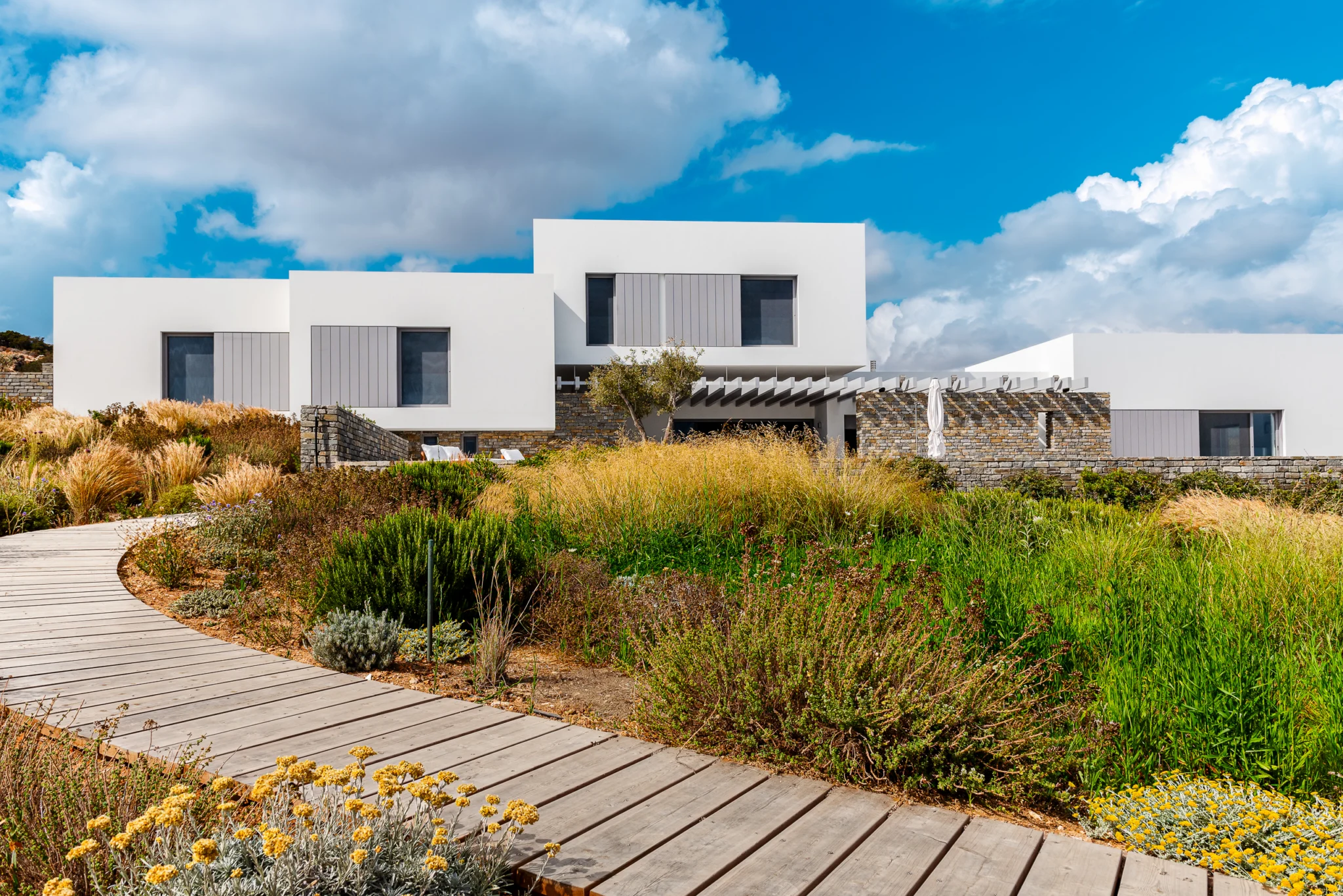 White cubic villa with grey shutters beside a garden boardwalk at Villa Seafront, Paros.