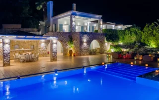 Elevated night view of Villa Stella, with illuminated stone arches, covered dining terraces and a blue-lit swimming pool in Porto Heli.