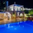 Elevated night view of Villa Stella, with illuminated stone arches, covered dining terraces and a blue-lit swimming pool in Porto Heli.