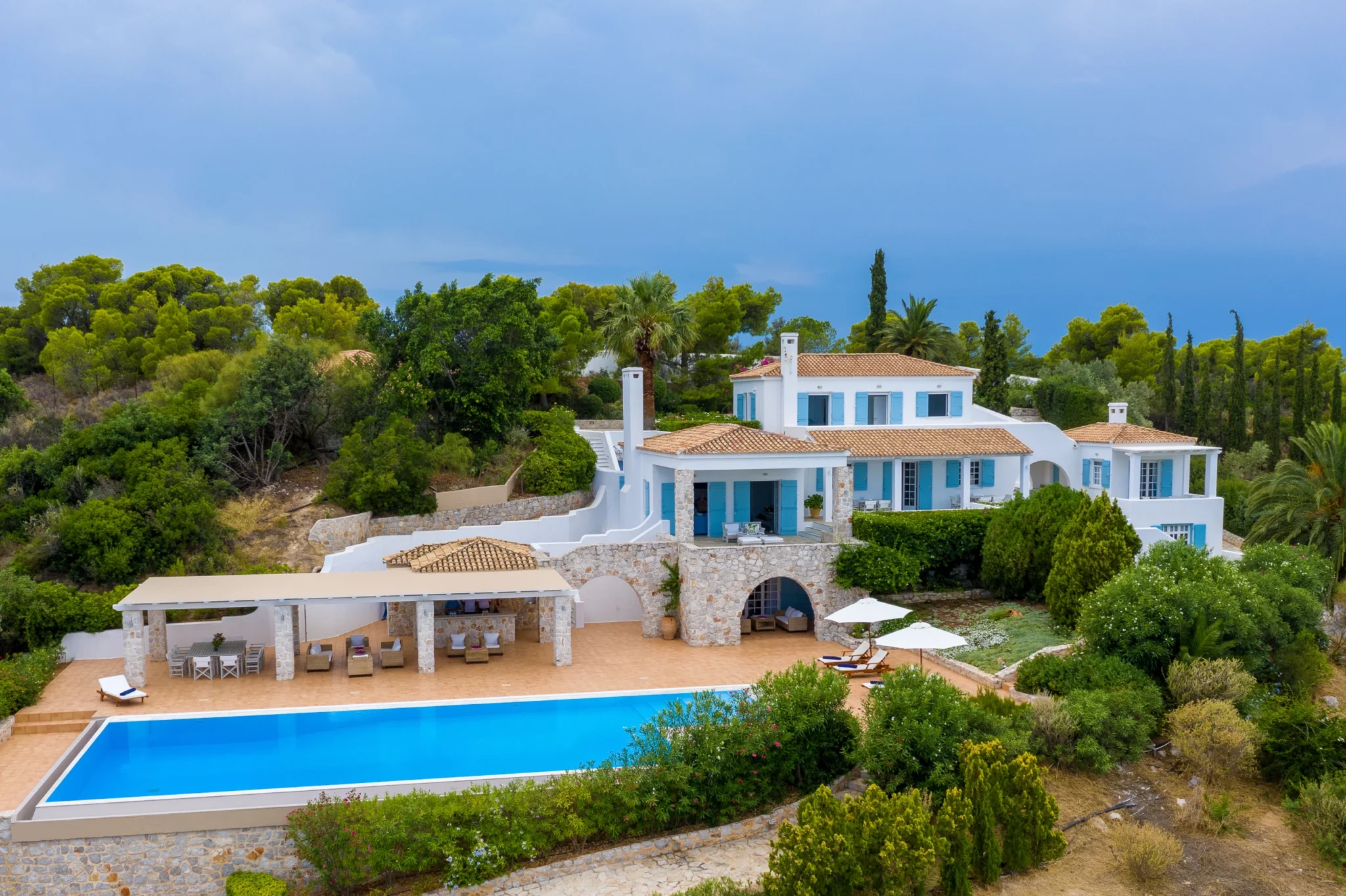 Aerial view of Villa Stella among trees, showing terracotta roofs, blue shutters, stone terraces and a long swimming pool in Porto Heli.