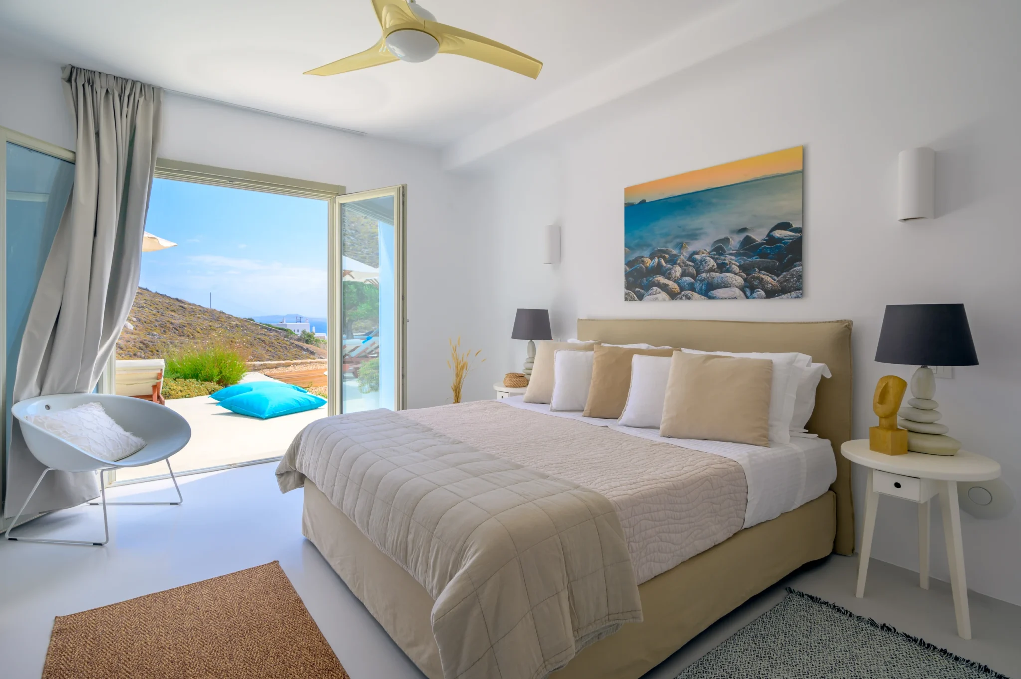 Bedroom with beige bedding, coastal artwork and open doors connecting the room to a terrace with turquoise cushions at Villa Velvet in Mykonos.