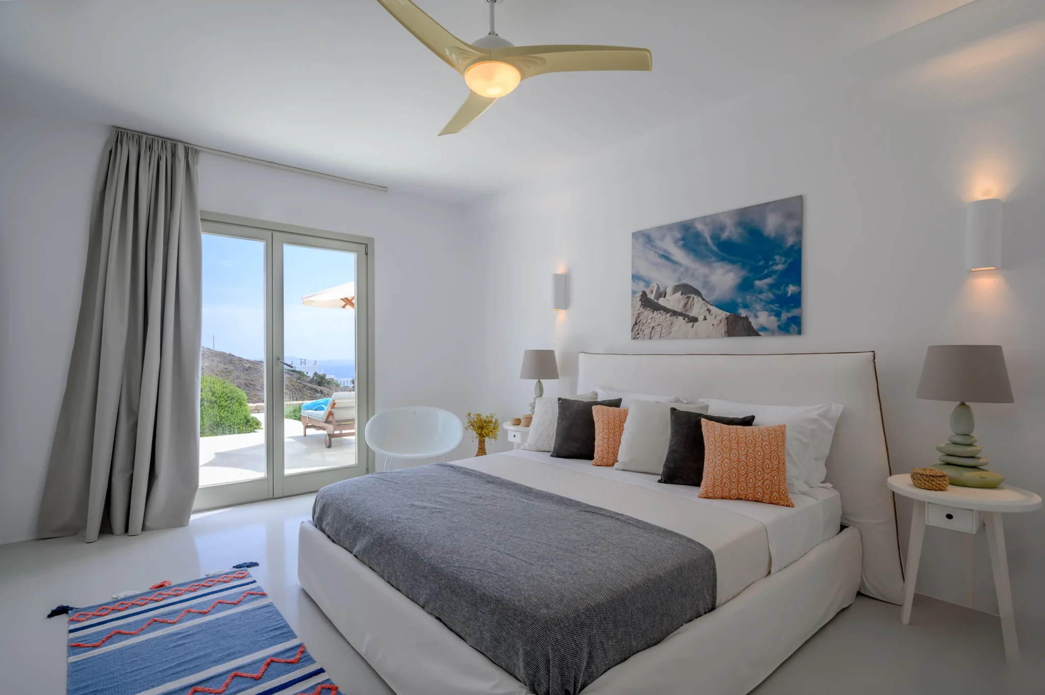 White bedroom with a grey bed throw, orange patterned cushions and glass doors opening onto the terrace at Villa Velvet in Mykonos.