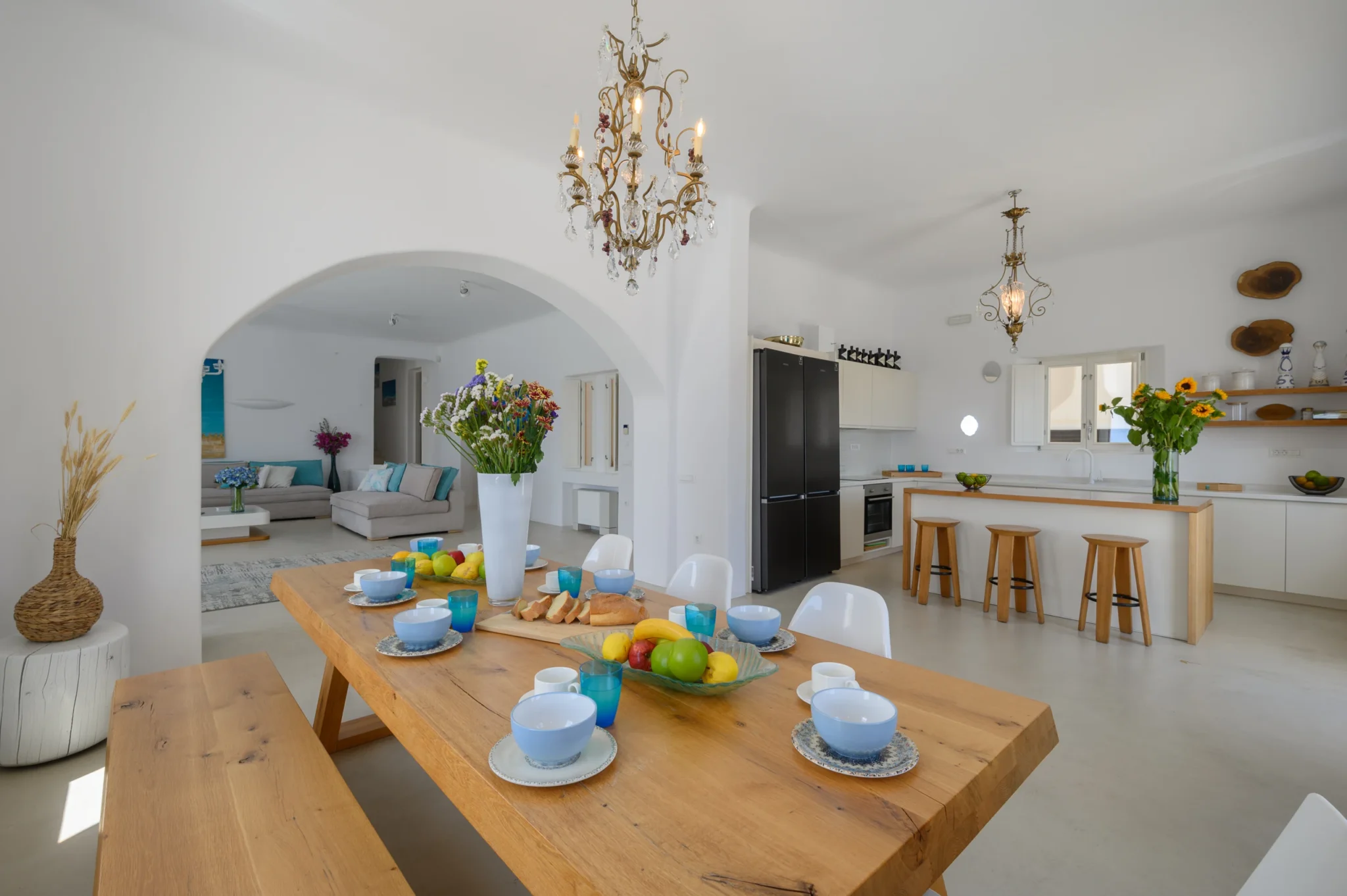 Timber breakfast table with blue bowls, fruit and flowers, leading towards the white kitchen and adjoining lounge at Villa Velvet in Mykonos.