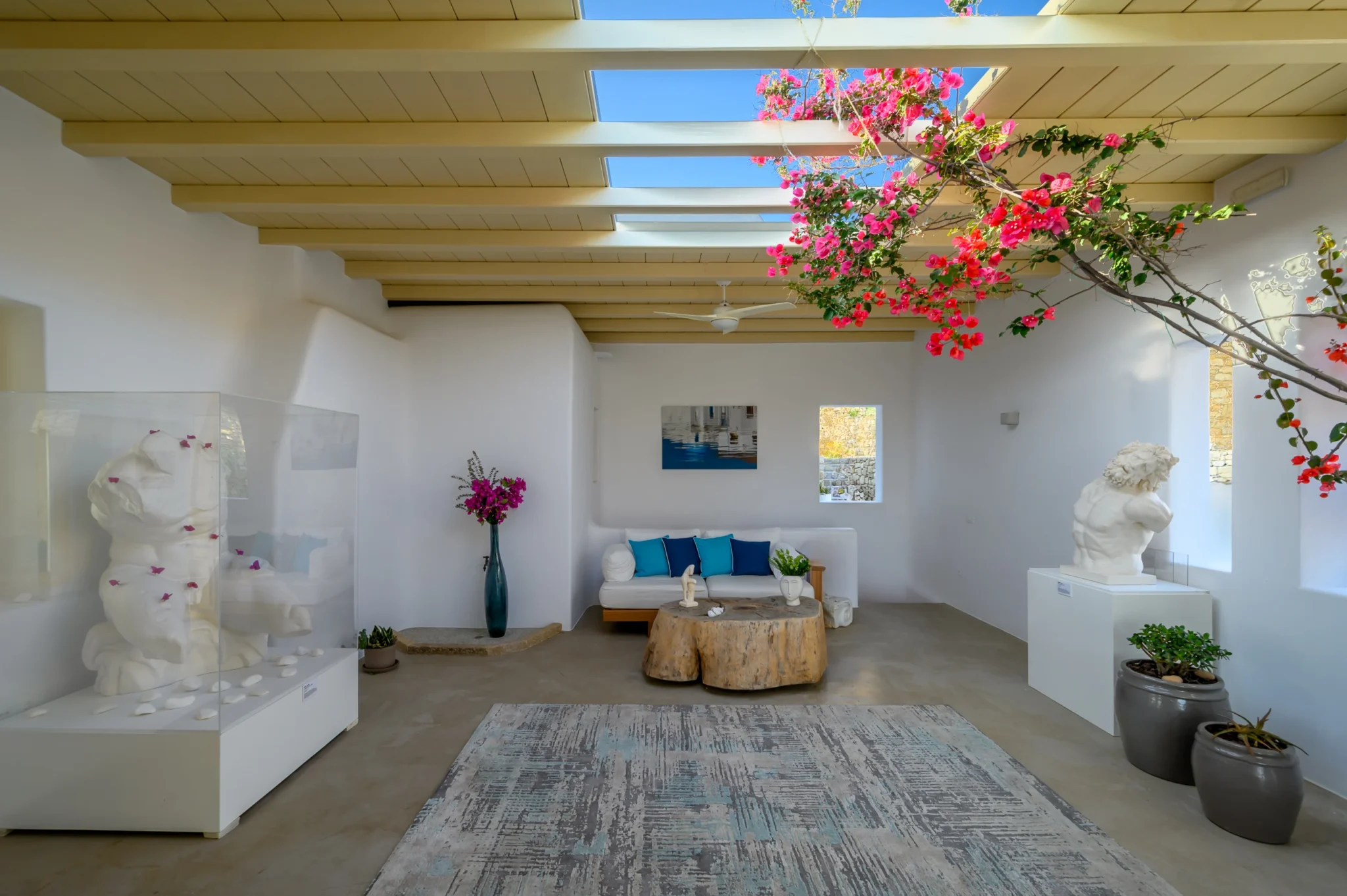 White courtyard lounge with blue cushions, sculptural figures and a low timber table beneath flowering bougainvillea and roof openings at Villa Velvet in Mykonos.