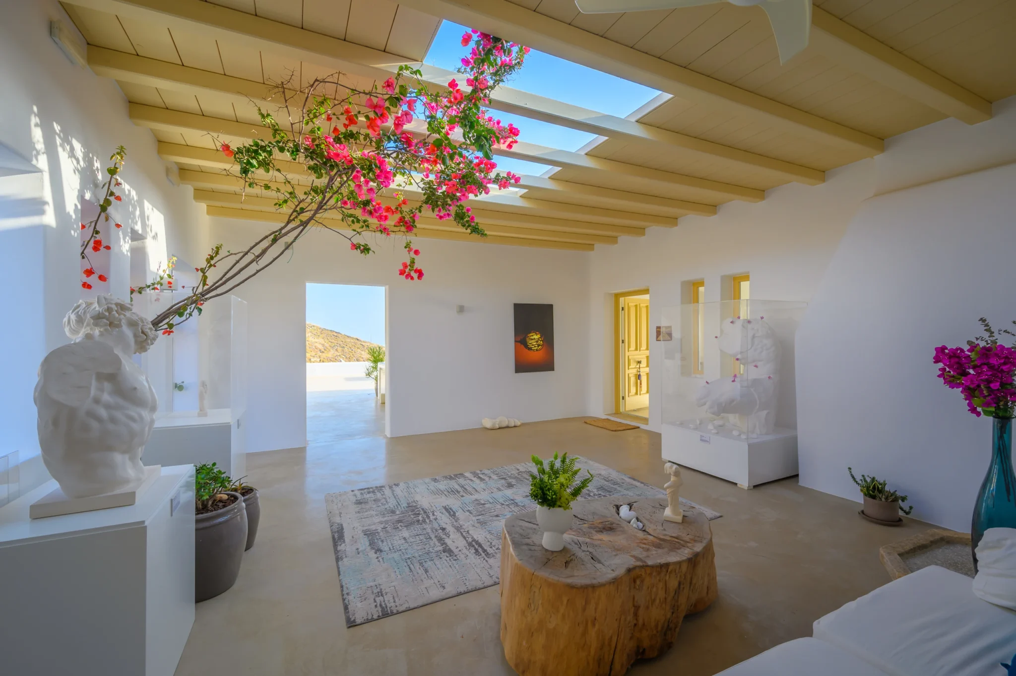 Sheltered white courtyard with sculptural figures, a timber coffee table and bougainvillea reaching through openings in the pergola roof at Villa Velvet in Mykonos.