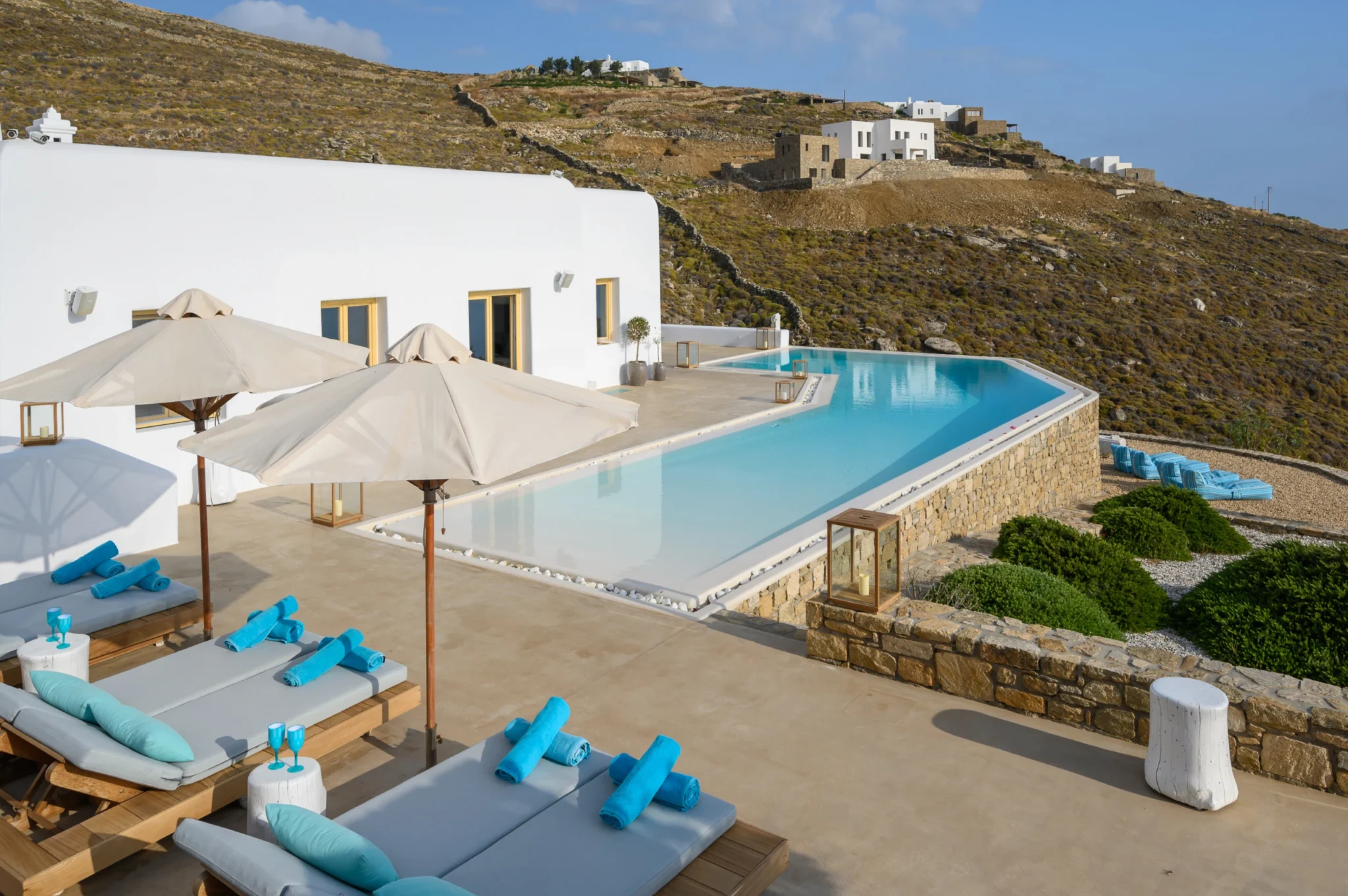 Curved swimming pool following the white villa facade, with parasols, loungers and planted stone terraces against the hillside at Villa Velvet in Mykonos.