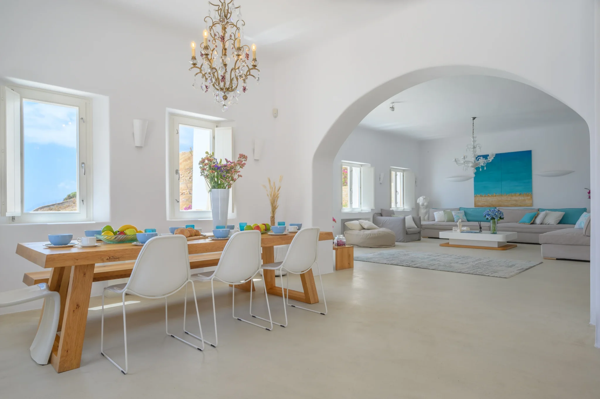 Breakfast table with white chairs beneath a chandelier, looking through a curved white arch into the living room at Villa Velvet in Mykonos.