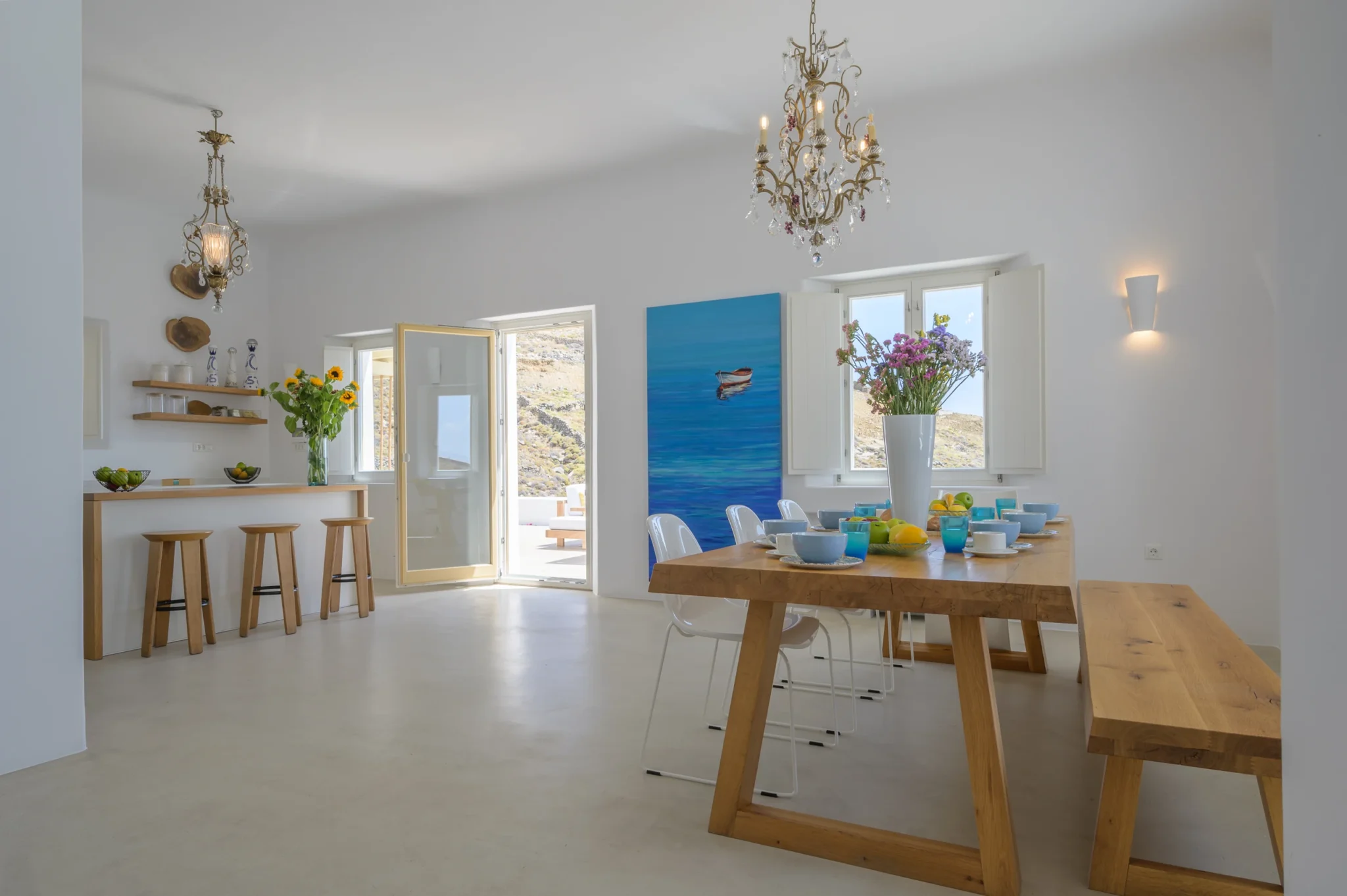 Timber dining table set for breakfast beside a white kitchen bar with three wooden stools and an open terrace door at Villa Velvet in Mykonos.