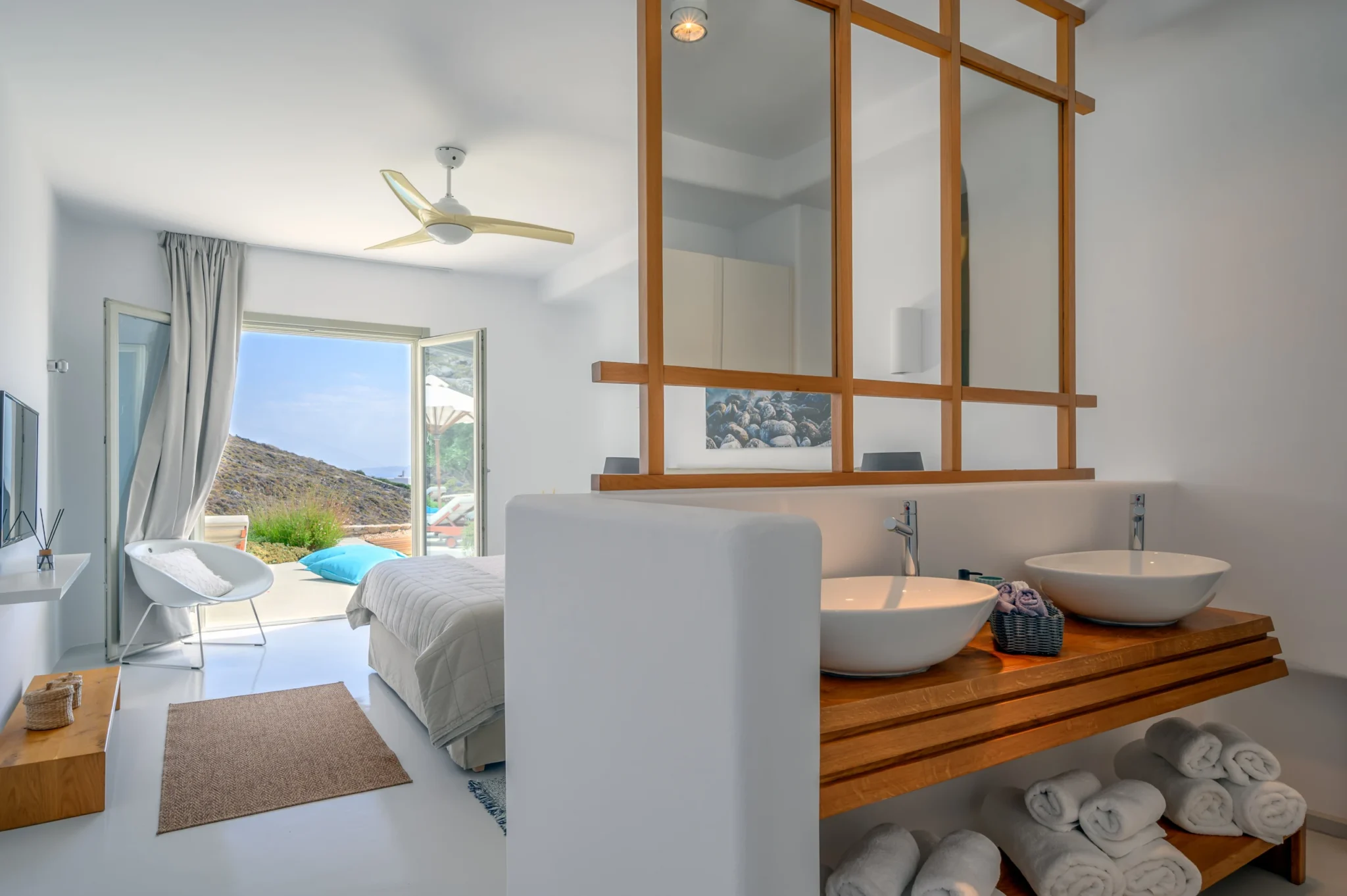 White double basins and timber vanity beside a bedroom with open glass doors to a sunlit terrace at Villa Velvet in Mykonos.
