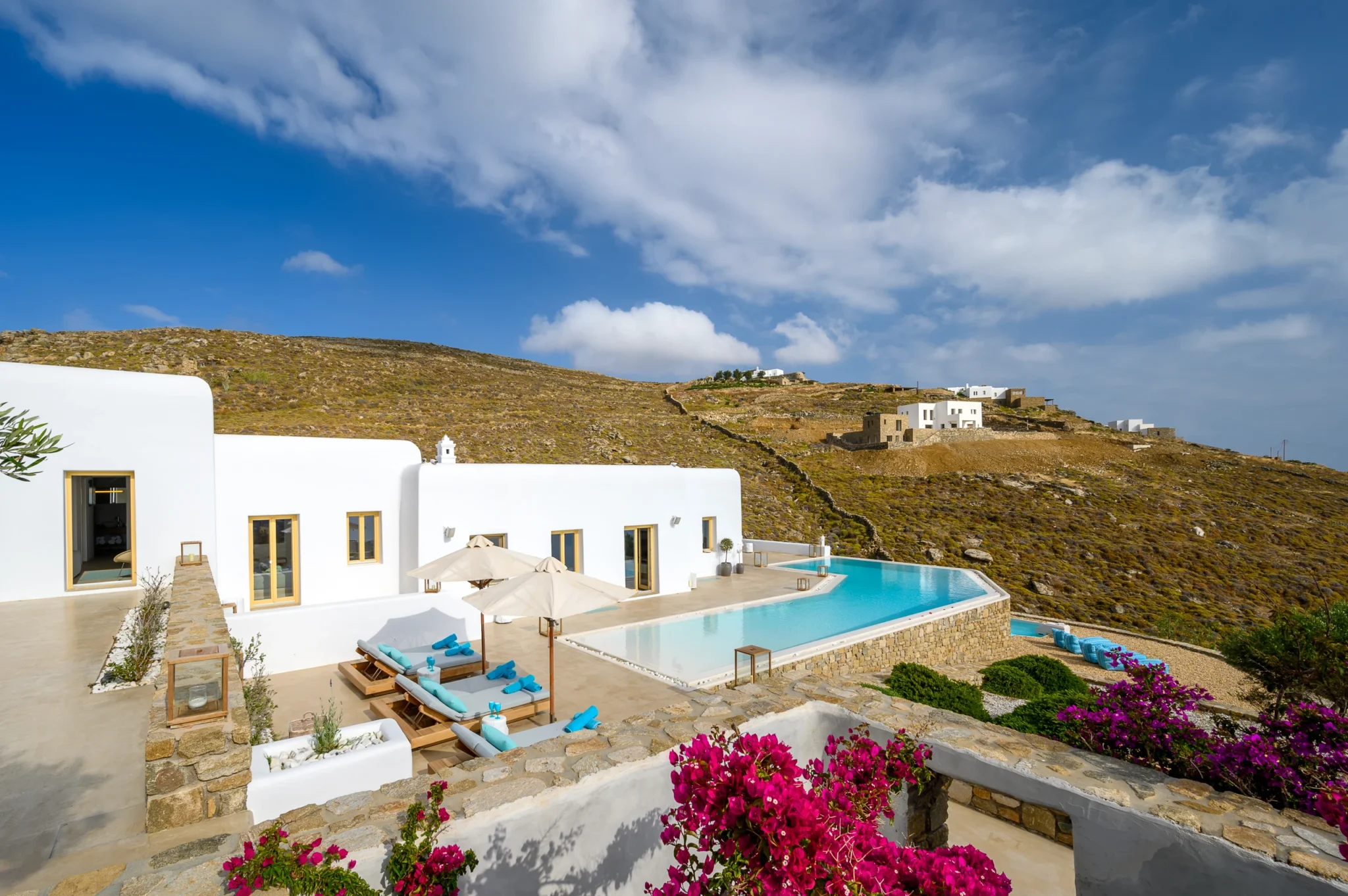 Elevated view of the white villa, curved swimming pool and sun loungers, framed by stone terraces and pink bougainvillea at Villa Velvet in Mykonos.