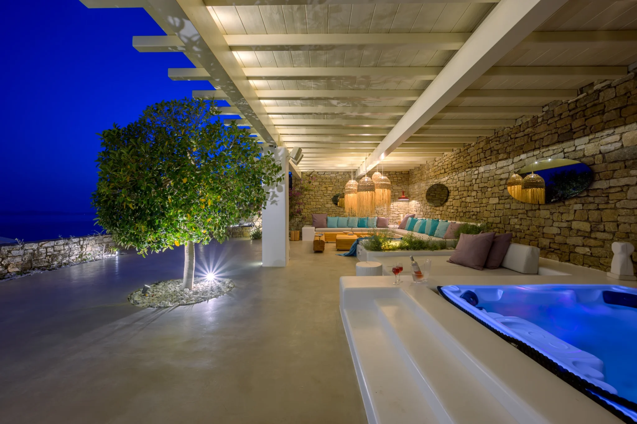 Blue-lit hot tub beside a long sheltered stone lounge with woven pendant lights and an illuminated tree at blue hour at Villa Velvet in Mykonos.