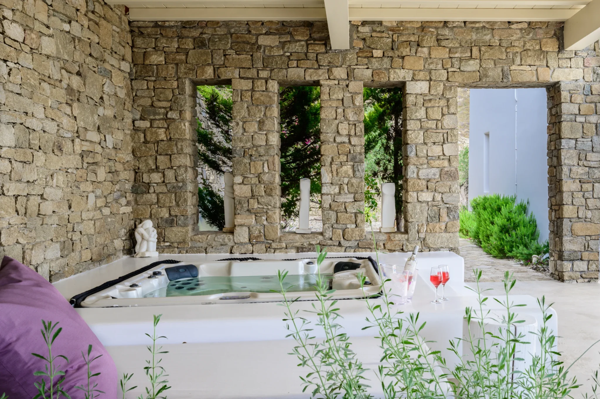 Hot tub beneath a stone pavilion with tall narrow openings framing garden greenery and a drinks setting beside the water at Villa Velvet in Mykonos.