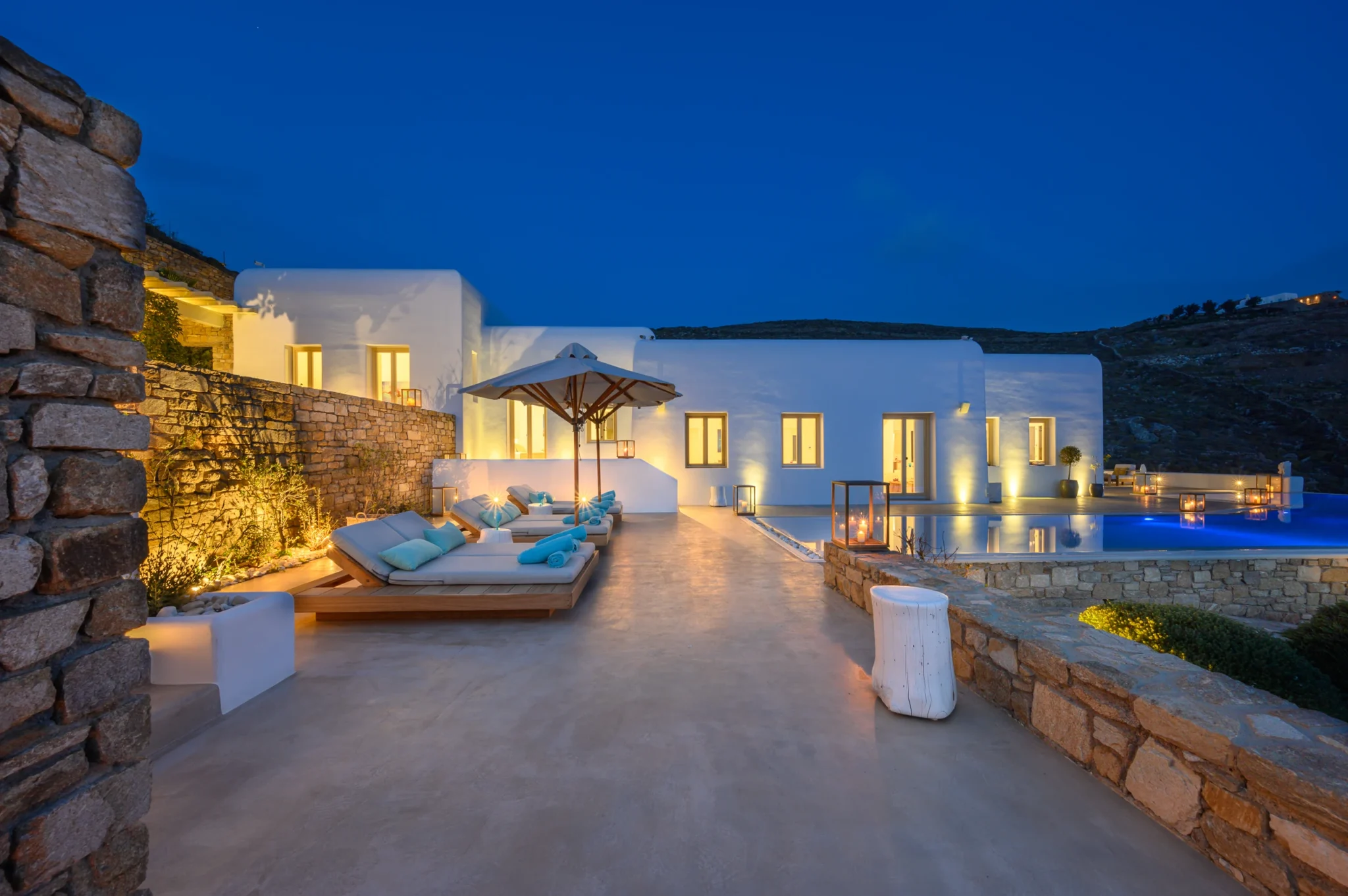 White villa and stone terraces illuminated at blue hour, with timber loungers and candle lanterns beside the blue swimming pool at Villa Velvet in Mykonos.