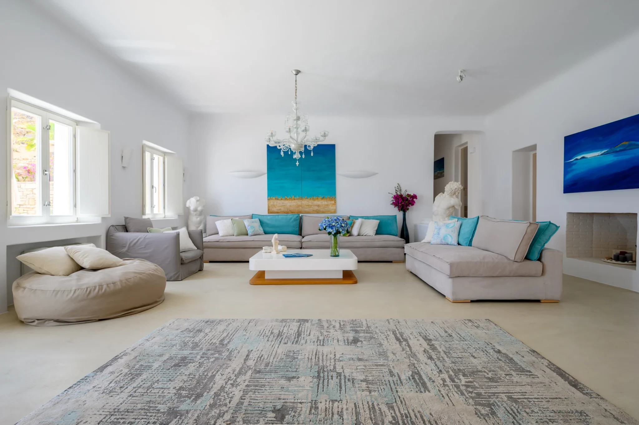 White living room with pale sofas, turquoise cushions, blue artwork, sculptural figures and a crystal chandelier at Villa Velvet in Mykonos.