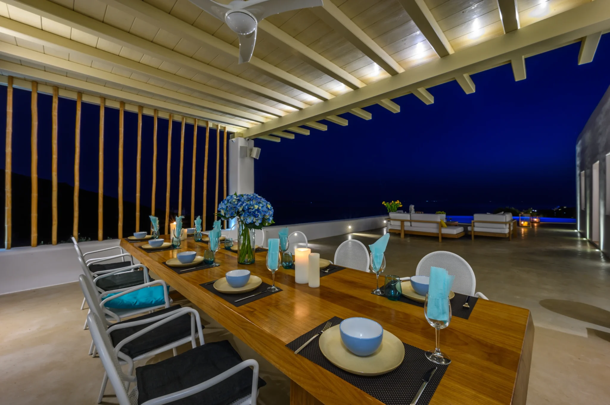 Timber dining table set with candles, turquoise napkins and blue hydrangeas beneath a pergola overlooking the pool terrace at night at Villa Velvet in Mykonos.