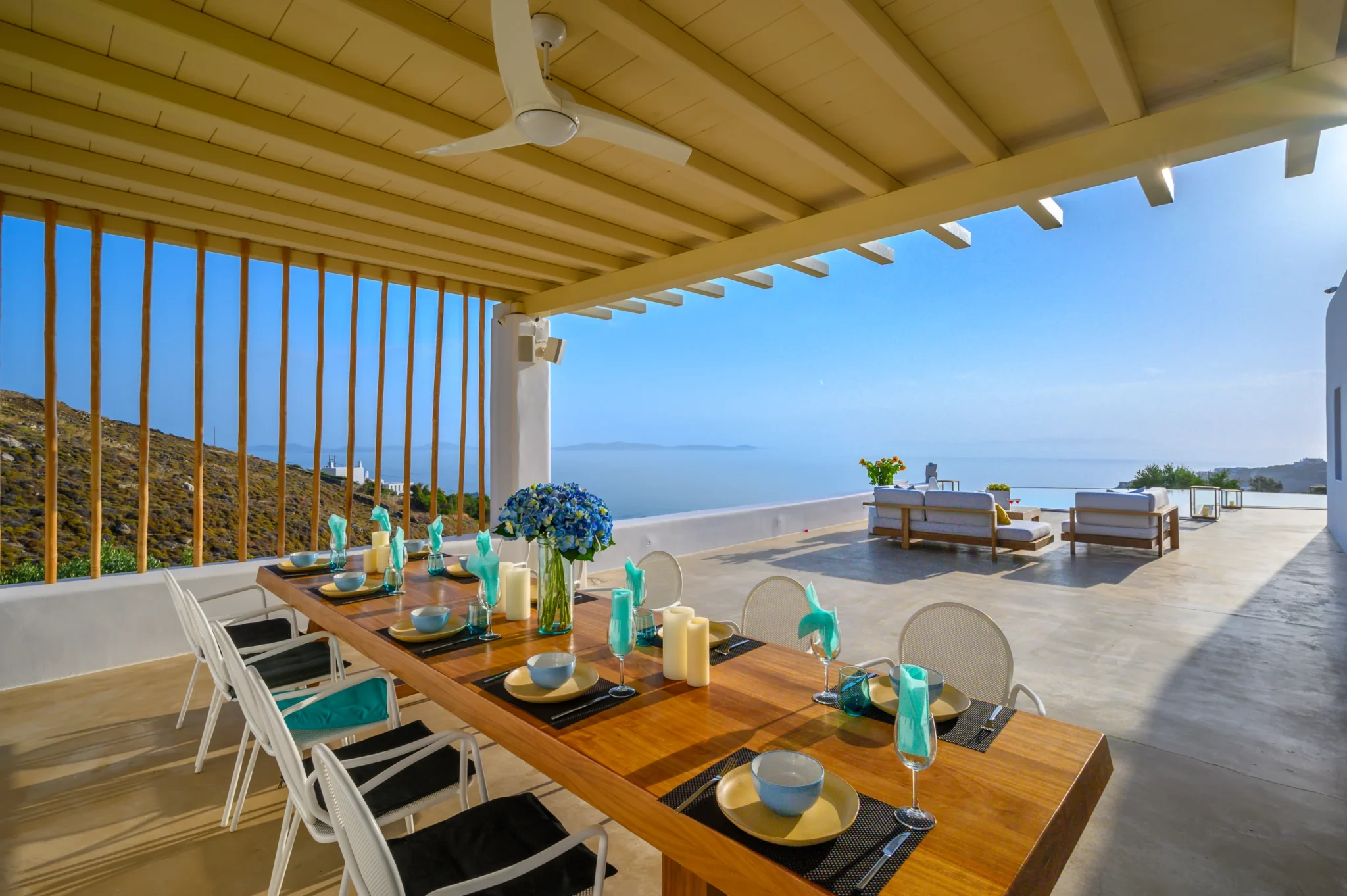 Set timber dining table beneath a pale pergola and vertical timber screen, looking towards the open lounge terrace and sea at Villa Velvet in Mykonos.