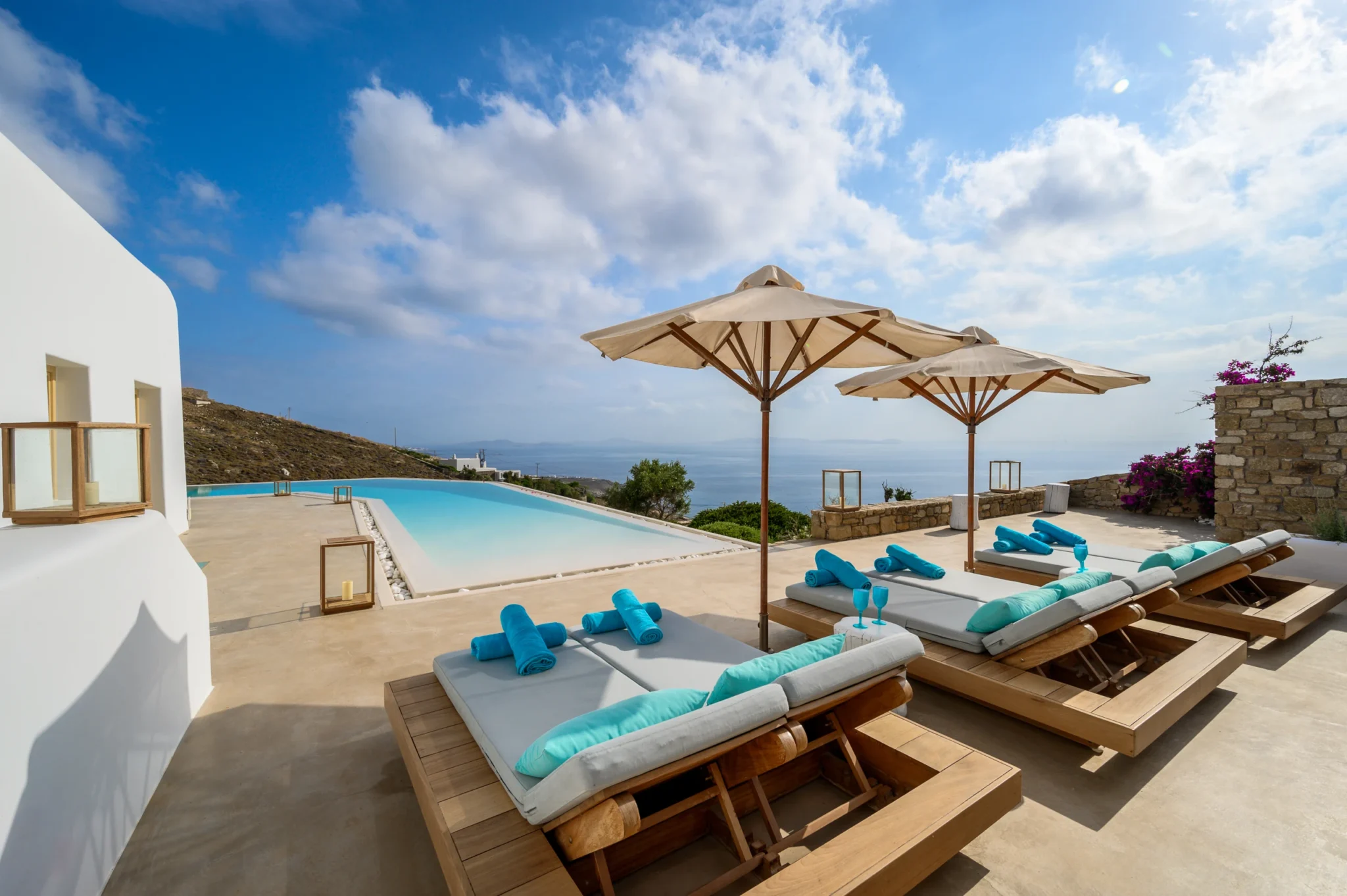 Timber sun loungers beneath two cream parasols beside the swimming pool, with the sea horizon beyond at Villa Velvet in Mykonos.