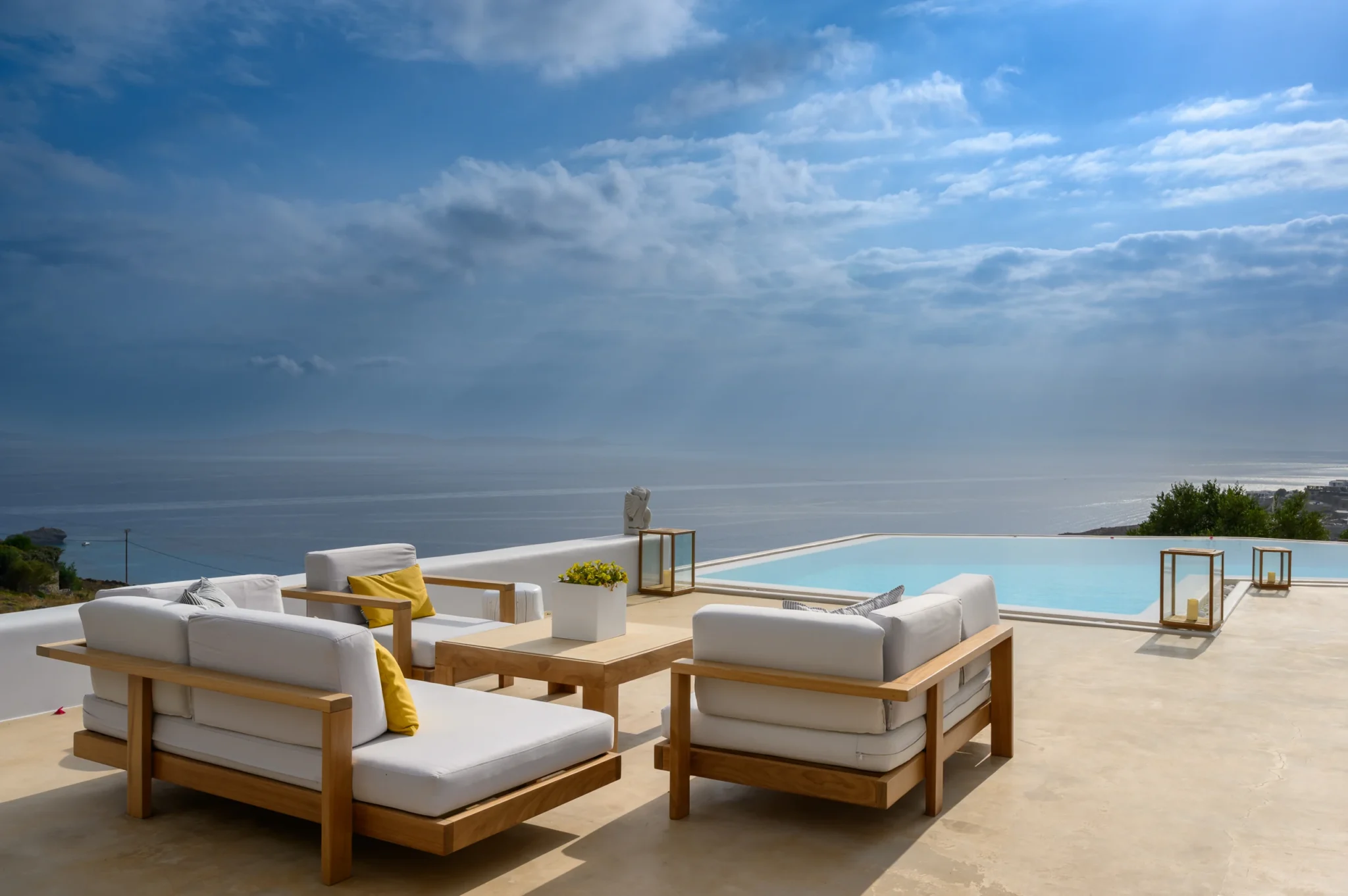 White-cushioned timber sofas with yellow accents on an open terrace beside the swimming pool, overlooking the sea at Villa Velvet in Mykonos.