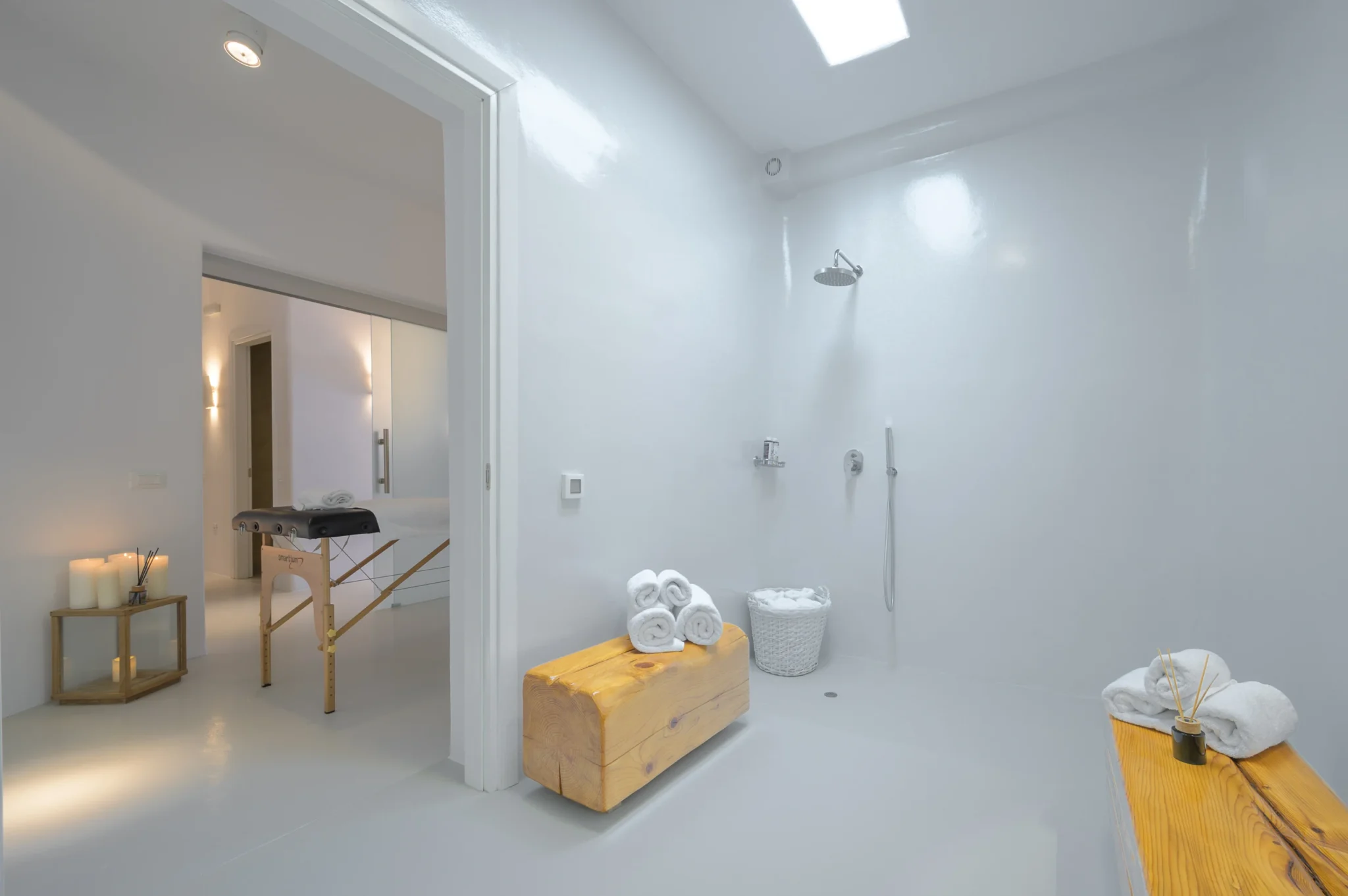 White shower room lit by a skylight, with timber benches and rolled towels beside a doorway to a massage table at Villa Velvet in Mykonos.