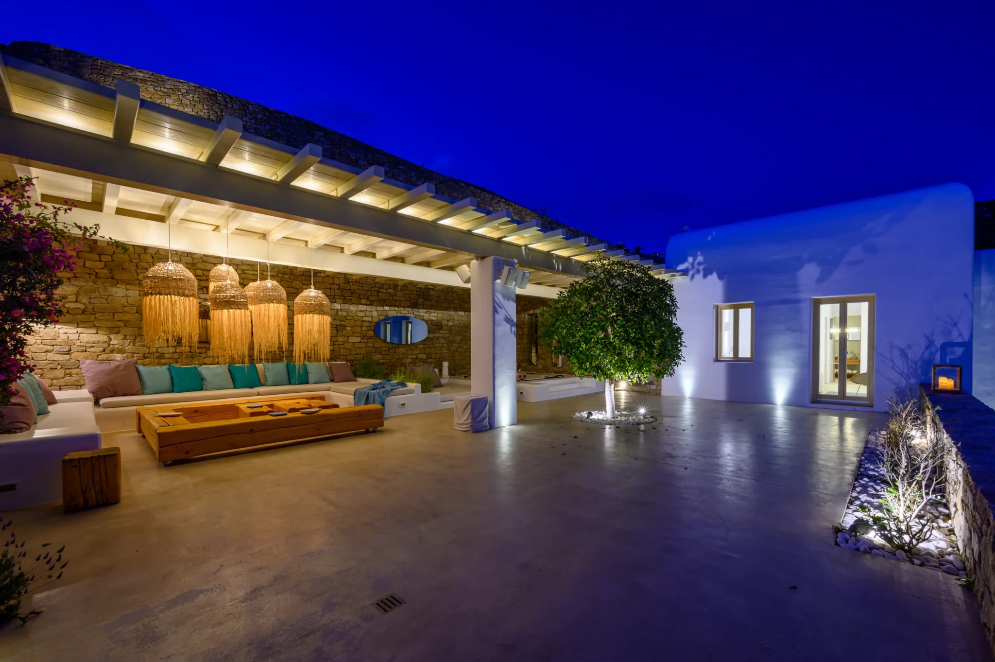 Covered stone lounge lit by fringed woven pendants beside an illuminated courtyard tree and white villa at blue hour at Villa Velvet in Mykonos.