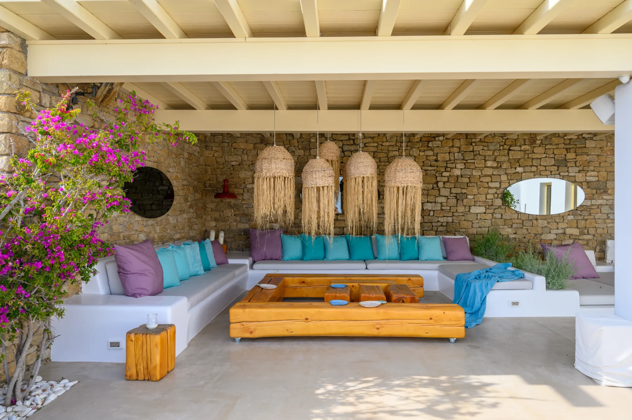 Sheltered stone lounge with built-in white seating, turquoise and purple cushions, fringed woven pendants and bougainvillea at Villa Velvet in Mykonos.