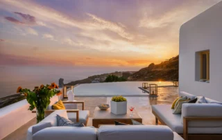 Sunset over the sea reflected in the swimming pool, with white-cushioned timber sofas, sunflowers and the white villa facade at Villa Velvet in Mykonos.