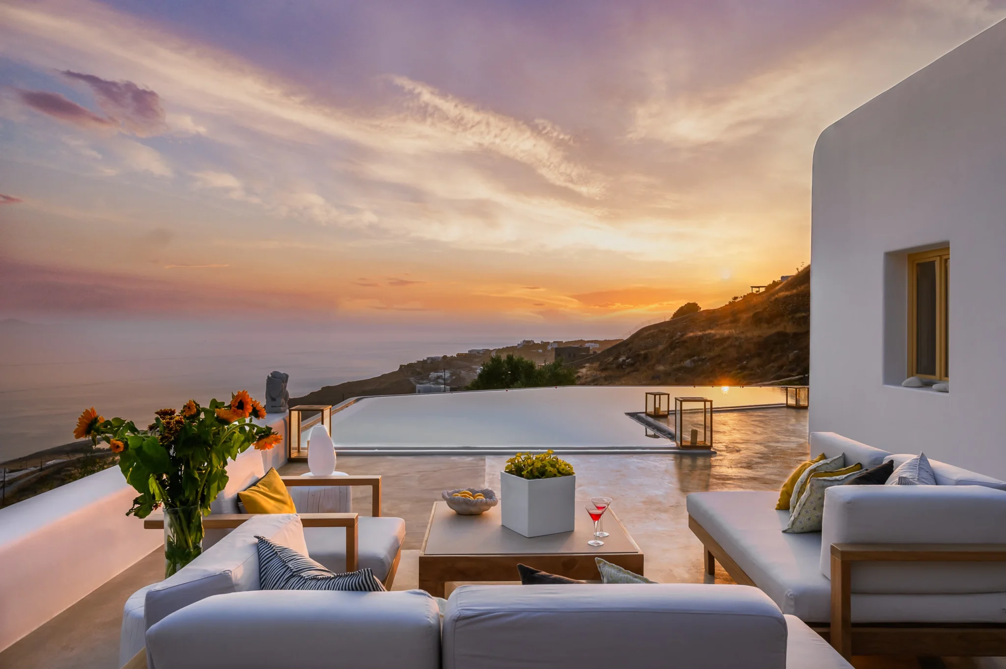 Sunset over the sea reflected in the swimming pool, with white-cushioned timber sofas, sunflowers and the white villa facade at Villa Velvet in Mykonos.