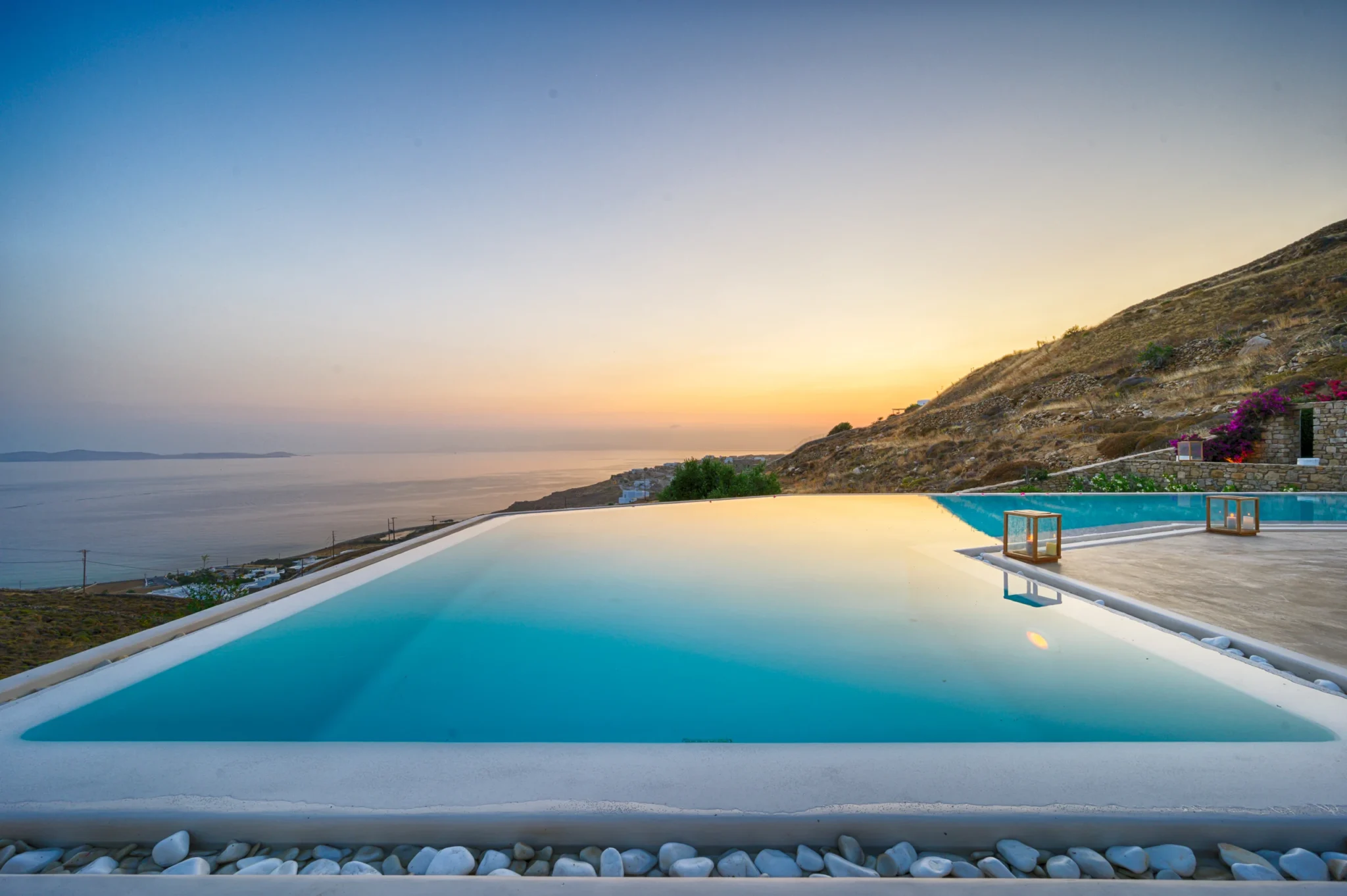 Still swimming pool reflecting the sunset sky, with timber lanterns along the terrace and the sea and hillside beyond at Villa Velvet in Mykonos.