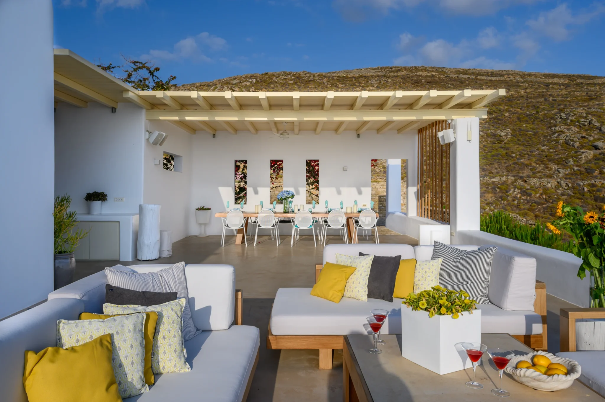 White-cushioned timber sofas with yellow accents and a flower-topped table leading towards the covered outdoor dining area at Villa Velvet in Mykonos.