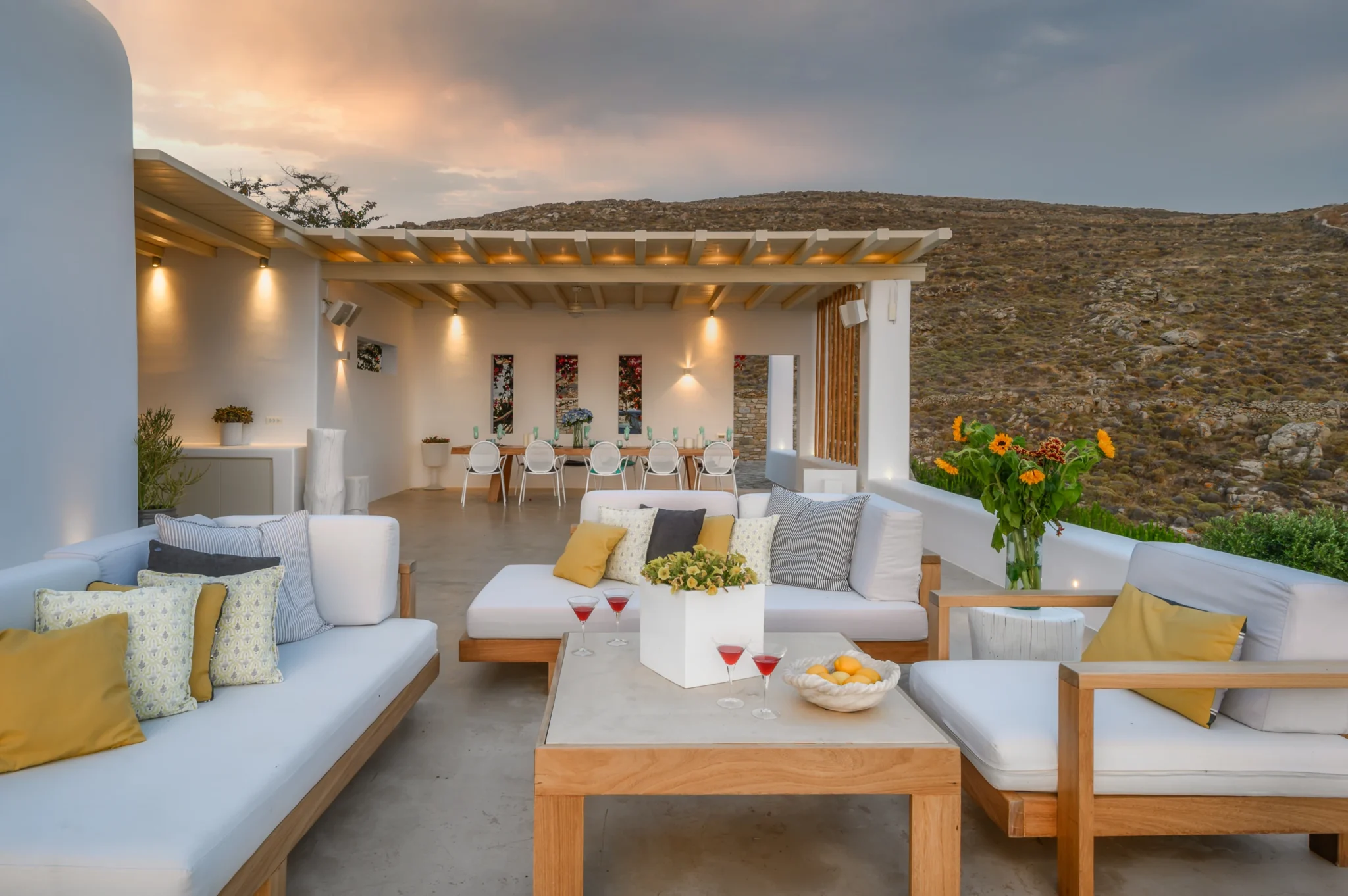 Outdoor sofas with yellow cushions in front of a pergola dining area illuminated by warm wall lights in the evening at Villa Velvet in Mykonos.
