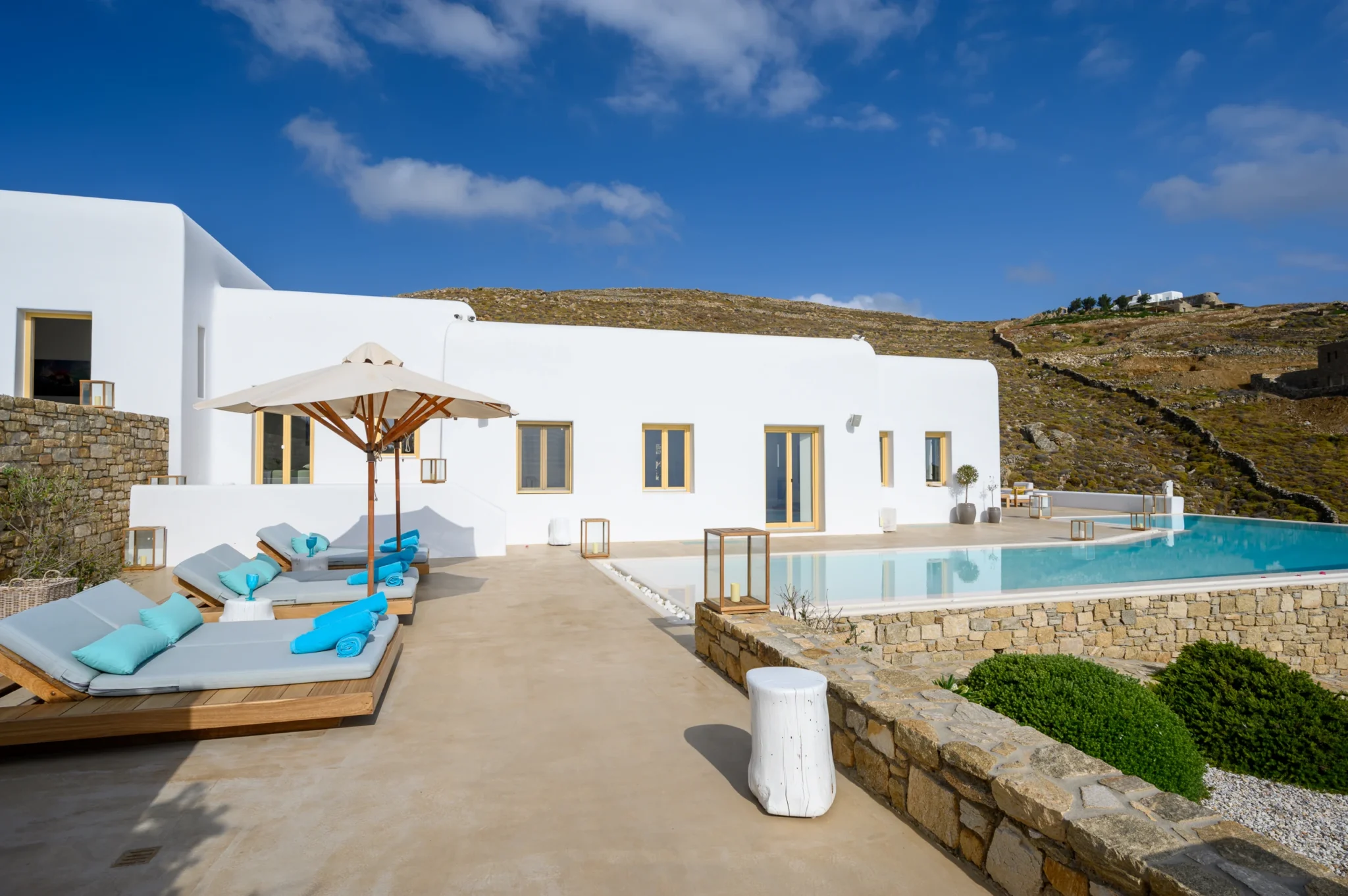 White Cycladic villa and swimming pool beside timber loungers with turquoise towels beneath a cream parasol at Villa Velvet in Mykonos.