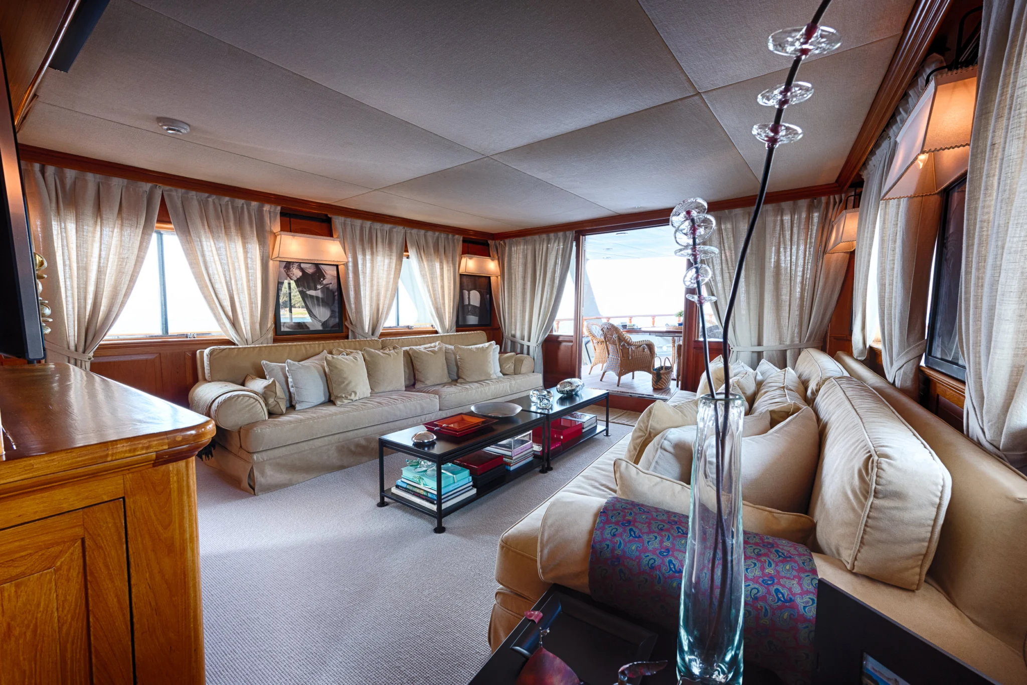 Walanka yacht saloon with timber cabinetry, pale sofas, curtains and an open doorway to the aft deck.