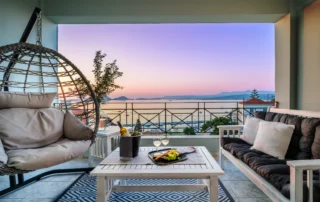 Balcony at dusk with a hanging chair, wine and a panoramic sea view at Zoe’s Sea View in Pylos, Greece.